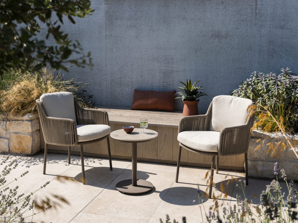 Zwei gepolsterte Serie Bernini von 4SO Dining-Stühle in Farbe Terre und ein kleiner runder Tisch stehen auf einem gefliesten Boden in einem sonnigen Garten. Eine Bank mit einem braunen Kissen und einer Topfpflanze ruht an einer Steinmauer, umgeben von üppigen Gräsern und Grünpflanzen.