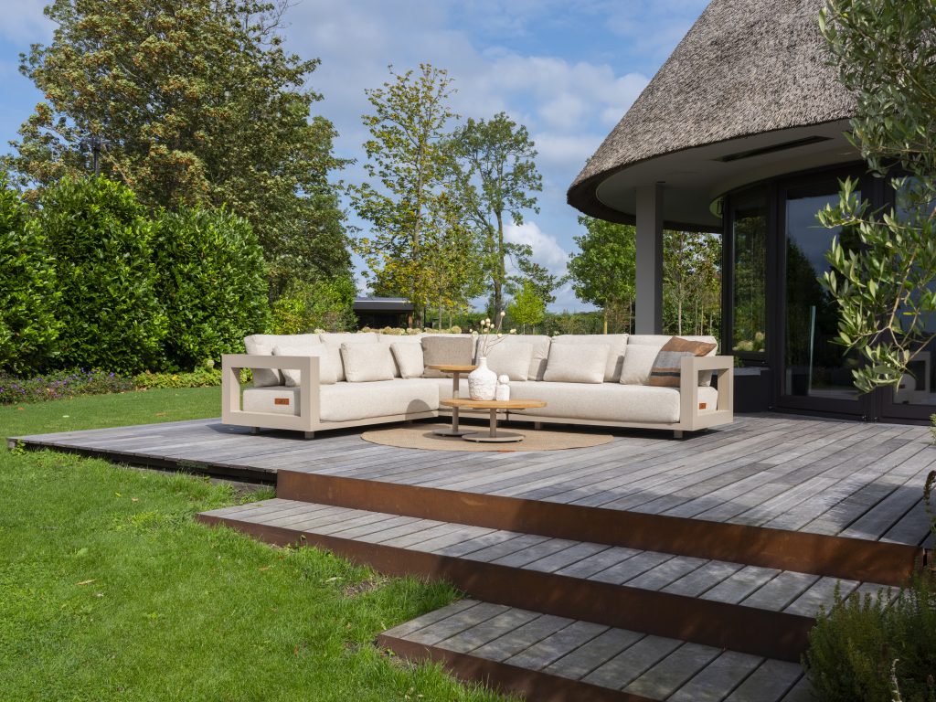 Ein großes Metropolitan Modular-Lounge-Set in Farbe Latte von 4SO mit Kissen sitzt auf einer Holzterrasse neben einem modernen Haus, umgeben von üppigem grünen Gras, Bäumen und Sträuchern unter einem teilweise bewölkten Himmel. Ein runder Volta-Tisch mit Dekoration steht vor dem Gartenmöbel.
