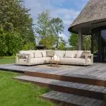 Ein großes Metropolitan Modular-Lounge-Set in Farbe Latte von 4SO mit Kissen sitzt auf einer Holzterrasse neben einem modernen Haus, umgeben von üppigem grünen Gras, Bäumen und Sträuchern unter einem teilweise bewölkten Himmel. Ein runder Volta-Tisch mit Dekoration steht vor dem Gartenmöbel.