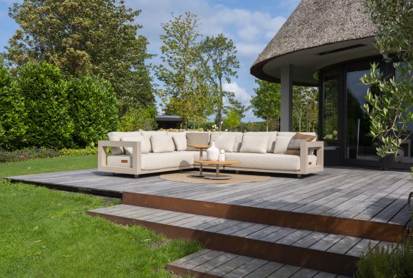 Ein großes Metropolitan Modular-Lounge-Set in Farbe Latte von 4SO mit Kissen sitzt auf einer Holzterrasse neben einem modernen Haus, umgeben von üppigem grünen Gras, Bäumen und Sträuchern unter einem teilweise bewölkten Himmel. Ein runder Volta-Tisch mit Dekoration steht vor dem Gartenmöbel.