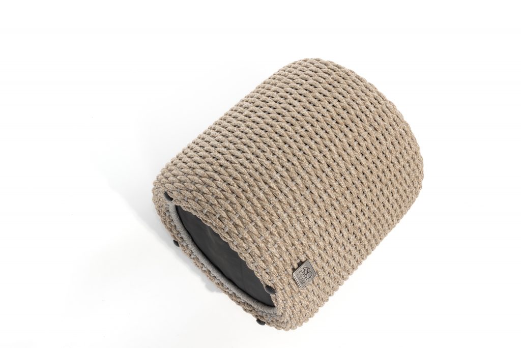 Ein zylindrischer Muffin-Sitzhocker von 4SO in Farbe Latte mit einem dicht gewebten beigen Seil außen, einem flachen schwarzen Sockel und einem kleinen Stoffanhänger ist eine stilvolle Ergänzung für Ihre Gartenmöbel vor einem strahlend weißen Hintergrund.