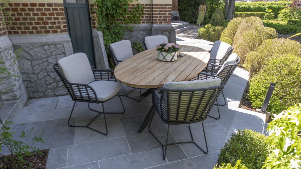 Ein Terrassenset aus der Gartenmöbel Serie Fabrice von 4SO mit einem ovalen Prado Holztisch und sechs gepolsterten Dining-Stuhl-Geflechtstühlen auf einem grauen Steinboden, umgeben von grünen Sträuchern und Ranken, die an einer Backsteinmauer emporranken.
