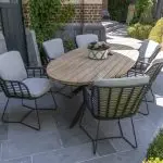 Ein Terrassenset aus der Gartenmöbel Serie Fabrice von 4SO mit einem ovalen Prado Holztisch und sechs gepolsterten Dining-Stuhl-Geflechtstühlen auf einem grauen Steinboden, umgeben von grünen Sträuchern und Ranken, die an einer Backsteinmauer emporranken.