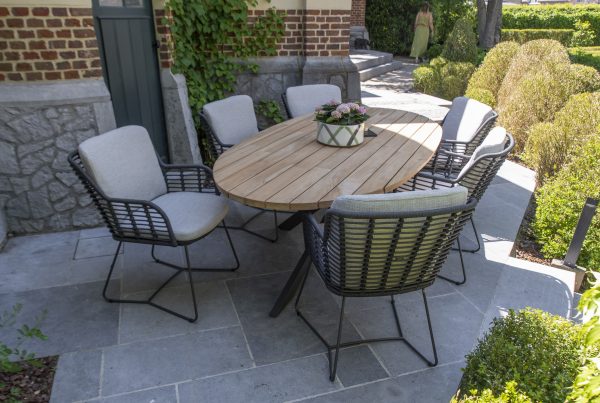 Ein Terrassenset aus der Gartenmöbel Serie Fabrice von 4SO mit einem ovalen Prado Holztisch und sechs gepolsterten Dining-Stuhl-Geflechtstühlen auf einem grauen Steinboden, umgeben von grünen Sträuchern und Ranken, die an einer Backsteinmauer emporranken.