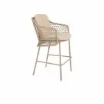 Ein moderner Barhocker aus der Serie Tramonti von 4SO in Latte mit geflochtener Rückenlehne und Seitenteilen im Rattanstil, gepolstertem Sitz und Rückenlehne sowie schlanken Metallbeinen mit Fußstütze - perfekt für stilvolle Gartenmöbel. Vor einem weißen Hintergrund.