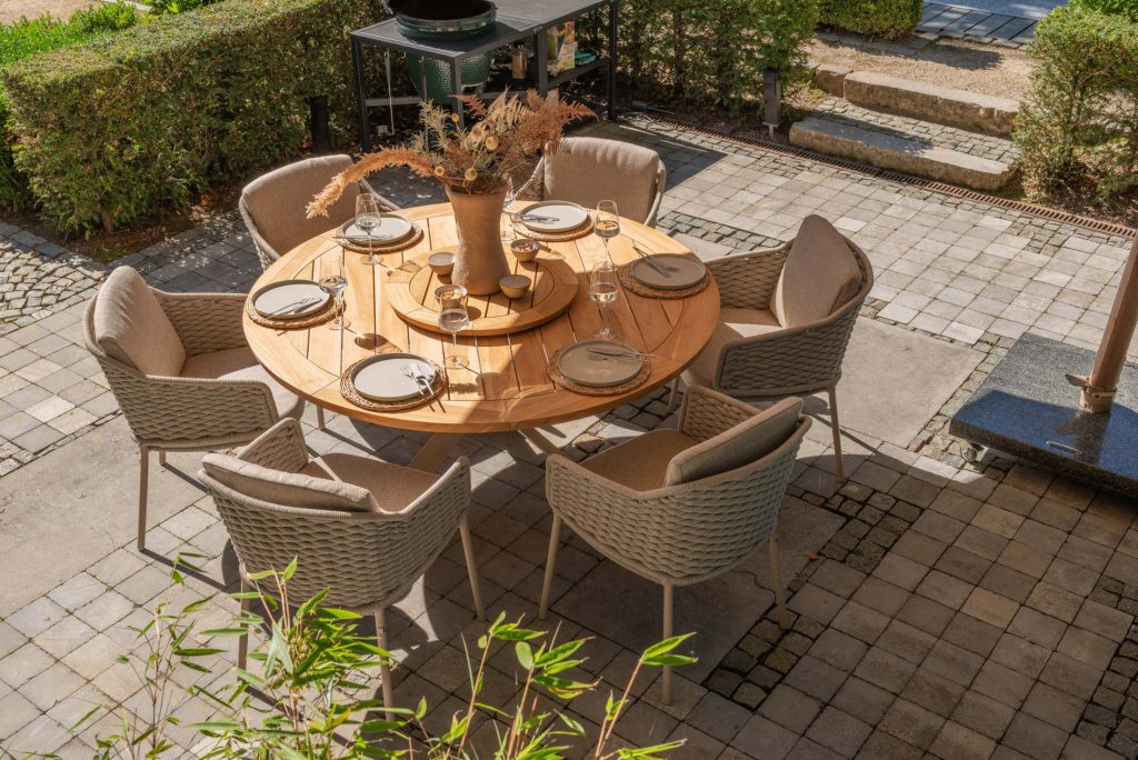 Ein runder Holztisch von Outdoor-Möbel ist für sechs Personen mit Tellern, Gläsern und Besteck gedeckt. Beige gepolsterte Stühle umgeben den Tisch. In der Mitte steht eine Vase mit getrockneten Gräsern. Die gepflasterte Terrasse ist von Hecken und Grünpflanzen umgeben.