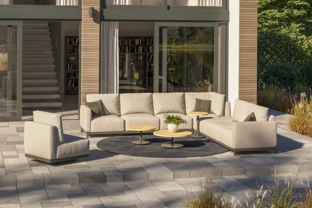Eine moderne Sitzecke auf der Terrasse mit beigen 4SO-Sectional-Sofas, einem passenden Drehstuhl und drei runden Couchtischen aus Holz auf einem grauen, runden Teppich. Die geflieste Terrasse verfügt über große Glastüren, vertikale Holzpaneele und eine üppige Aussicht - der perfekte Gartenmöbel-Stil.