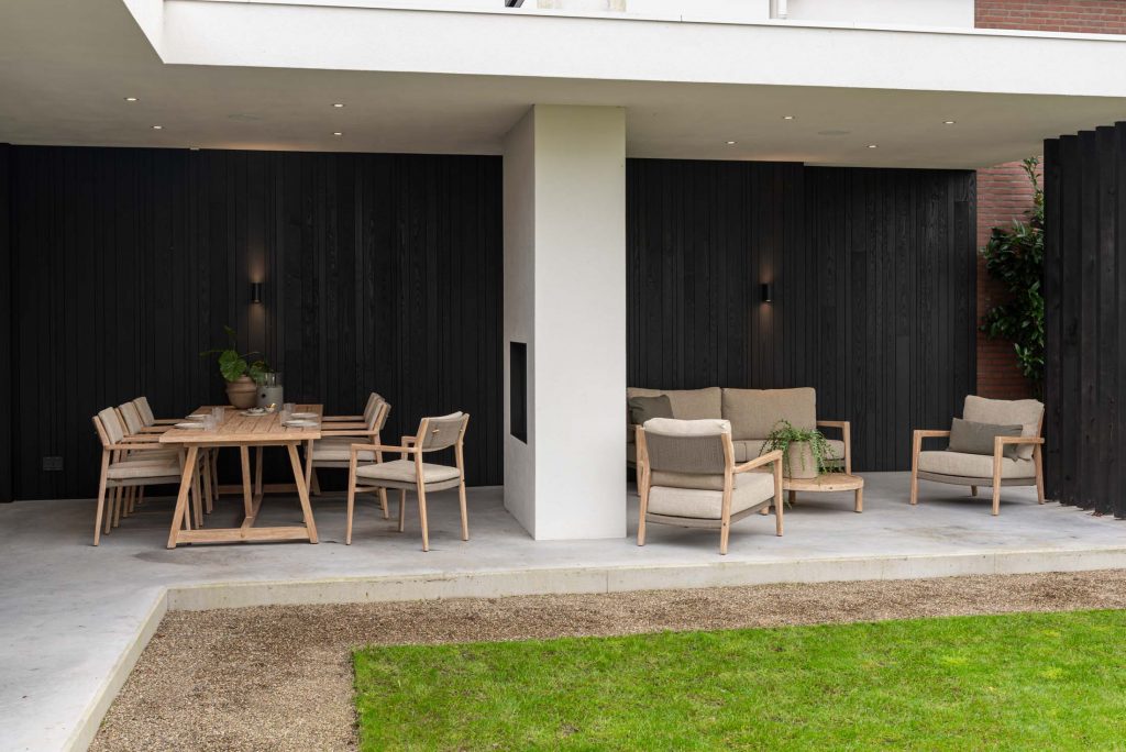 Eine moderne überdachte Terrasse mit 4SO Outdoor-Möbel: links ein Esstisch aus Holz für sechs Personen und rechts ein Lounge-Bereich mit Sofas und Sesseln. Betonböden, schwarze Holzwände, sanfte Beleuchtung und ein grüner Rasen vervollständigen den einladenden Raum.