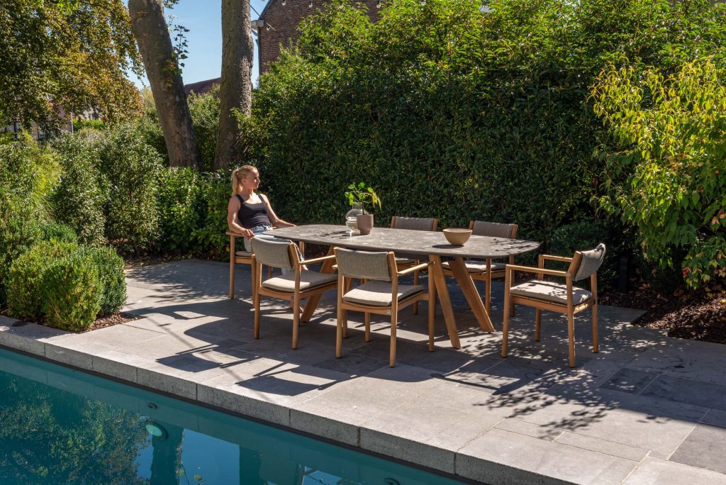 Eine Frau in einem schwarzen Kleid sitzt allein an einem großen Outdoor-Möbel-Esstisch mit acht Holzstühlen neben einem Swimmingpool, umgeben von üppig grünen Bäumen und Büschen an einem sonnigen Tag in Hamburg. Eine Vase mit grünen Zweigen schmückt den Tisch.