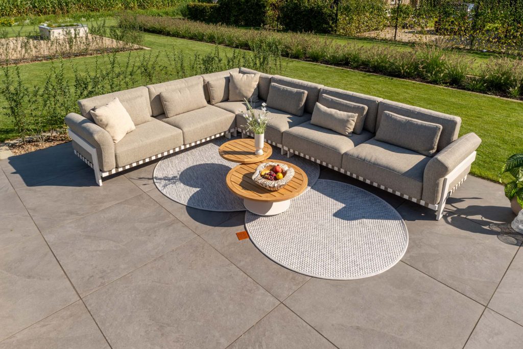 Ein beigefarbenes Outdoor-Sectional-Sofa mit Kissen steht auf einer Terrasse mit zwei runden Holztischen auf weißen Teppichen. Auf diesen stilvollen Gartenmöbeln stehen eine Vase mit Blumen und eine Schale mit Obst, mit Blick auf einen angelegten Garten und Rasen.