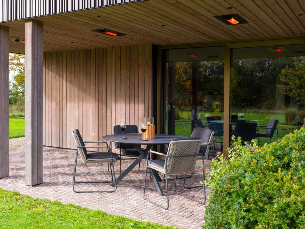 Auf einer modernen Terrasse von 4SO stehen ein runder schwarzer Tisch, vier Stühle und ein Glas Wein. Im Schatten eines hölzernen Überhangs und umgeben von Grün, sitzt das Gartenmöbel-Set neben großen Glastüren, die den Garten widerspiegeln.