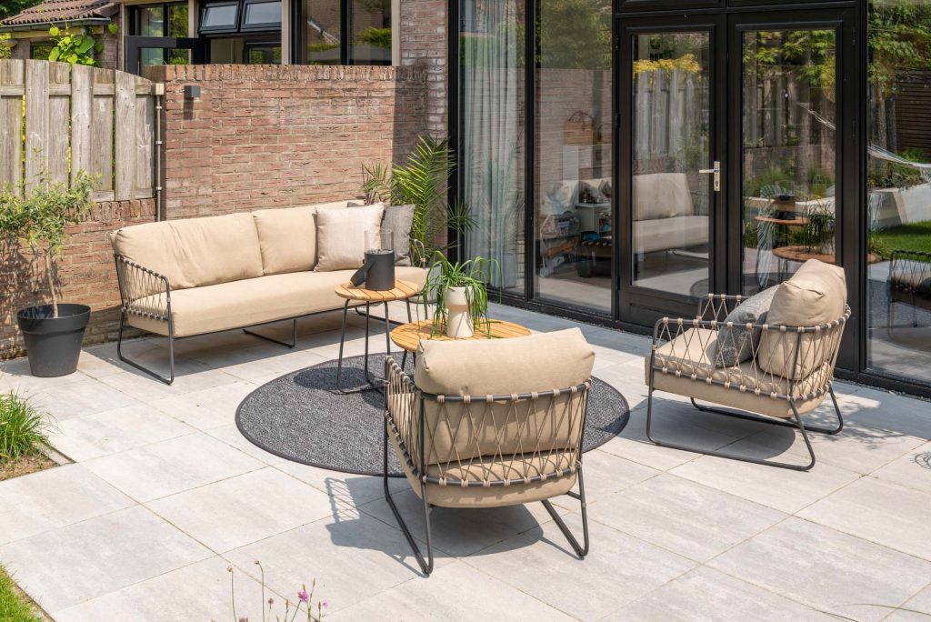 Terrasse im Freien mit beigem Polstersofa 4SO, passenden Sesseln und runden Holztischen auf grauem Teppich. Glastüren spiegeln den Garten wider; Pflanzen und ein Holzzaun begrenzen dieses gepflasterte, von Hamburg inspirierte Geschäft.
