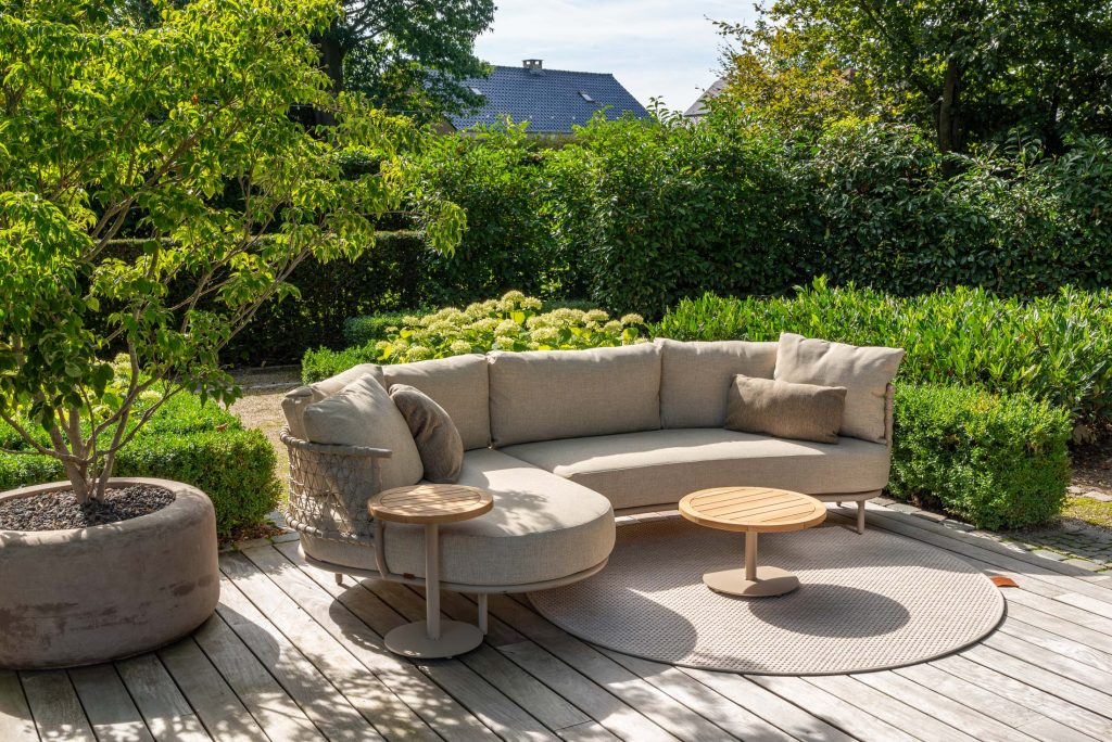 Eine moderne Außenterrasse in Hamburg mit einem geschwungenen beigen Outdoor-Möbel-Sofa, passenden Kissen, zwei runden Holztischen und einer großen Topfpflanze auf einem Holzdeck, umgeben von üppigem Grün und Hecken.