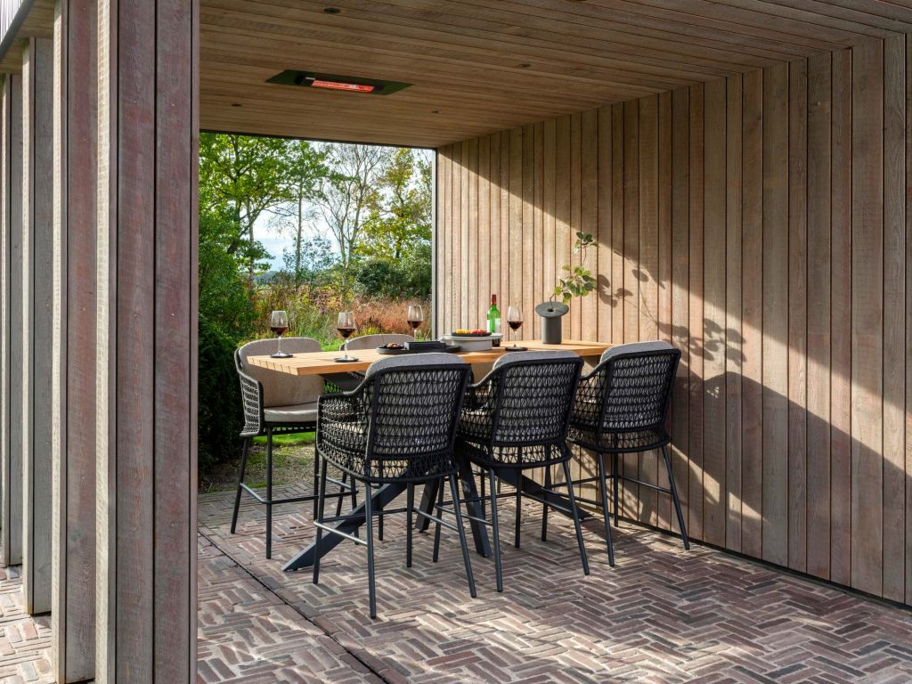 Ein moderner Essbereich im Freien mit Gartenmöbeln, einem Dach und Wänden aus Holz, einem rechteckigen Tisch für vier Personen mit Weingläsern und Flaschen, umgeben von schwarzen geflochtenen Stühlen, mit Blick auf einen Garten.