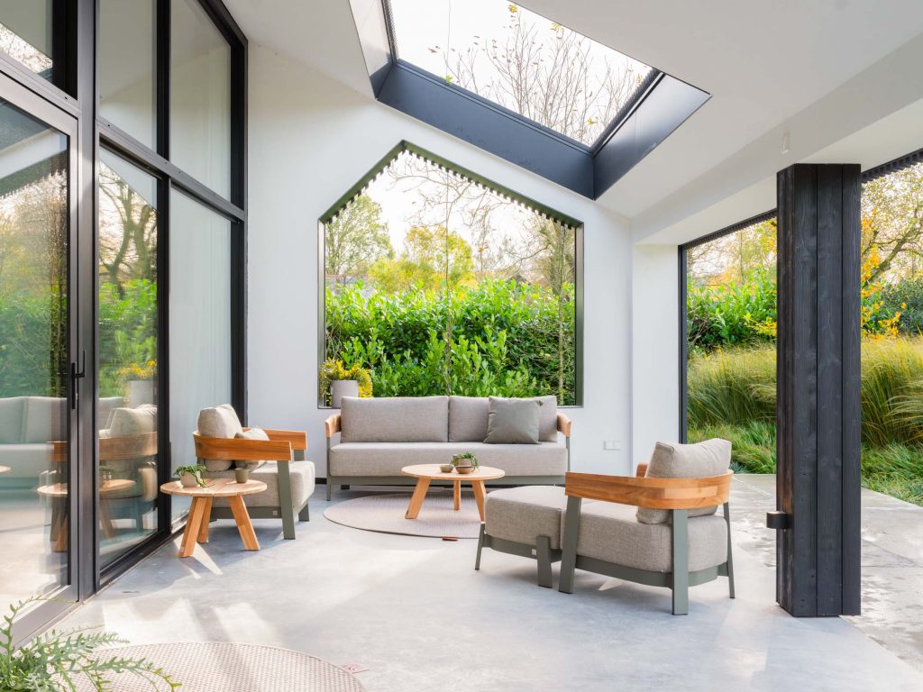 Ein moderner Wintergarten mit großen Fenstern, einem Oberlicht und neutralfarbenen 4SO-Sofas schafft einen bequemen Sitzbereich. Hölzerne Akzente und Grünpflanzen im Außenbereich füllen diesen von Hamburg inspirierten Raum mit natürlichem Licht und sorgen für eine luftige Geschäftsatmosphäre.