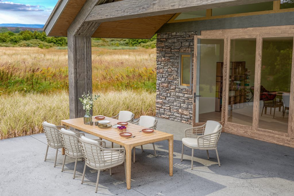 Ein moderner Essbereich im Freien mit einem hellen Liam Holztisch für sechs Personen und Dining-Stühle der Serie Como von 4SO. Die Gartenmöbel stehen neben Steinmauern und Glastüren, mit Blick auf eine Wiese unter blauem Himmel.
