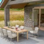 Ein moderner Essbereich im Freien mit einem hellen Liam Holztisch für sechs Personen und Dining-Stühle der Serie Como von 4SO. Die Gartenmöbel stehen neben Steinmauern und Glastüren, mit Blick auf eine Wiese unter blauem Himmel.