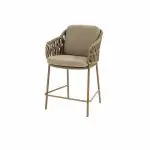 Der High-Dining-Stuhl der Serie Biarritz verfügt über beigefarbene Sitz- und Rückenkissen, einen hellbraunen Rahmen aus geflochtenem Rattan mit Armlehnen und schlanke Metallbeine mit einer Fußstütze. Der Hintergrund ist einfarbig weiß.