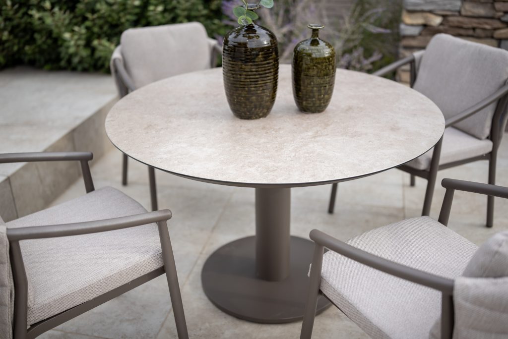 Ein runder Sarah Diningtisch 130cm von 4SO in Keramikdruck Terre mit einem stabilen Sockel ist umgeben von vier gepolsterten, Barolo Dining-Stühlen von 4SO. Zwei grüne Vasen mit Struktur stehen auf dem Tisch auf einer Terrasse mit Steinfliesen und Grünpflanzen.