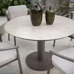 Ein runder Sarah Diningtisch 130cm von 4SO in Keramikdruck Terre mit einem stabilen Sockel ist umgeben von vier gepolsterten, Barolo Dining-Stühlen von 4SO. Zwei grüne Vasen mit Struktur stehen auf dem Tisch auf einer Terrasse mit Steinfliesen und Grünpflanzen.