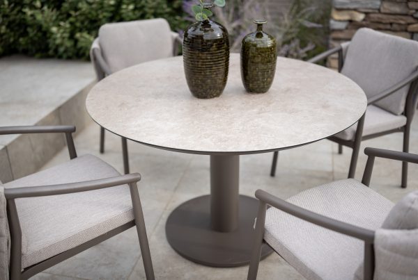 Ein runder Sarah Diningtisch 130cm von 4SO in Keramikdruck Terre mit einem stabilen Sockel ist umgeben von vier gepolsterten, Barolo Dining-Stühlen von 4SO. Zwei grüne Vasen mit Struktur stehen auf dem Tisch auf einer Terrasse mit Steinfliesen und Grünpflanzen.