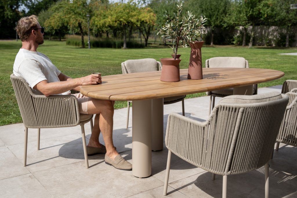 Ein Mann mit Sonnenbrille und Freizeitkleidung sitzt an einem Estate Gartenmöbel-Diningtisch teak natur von 4SO mit sechs beigen Bernini Stühlen auf einer Veranda. Auf dem Tisch in Mangoform stehen zwei Terrakotta-Vasen mit Pflanzen. Grünes Gras und Sonnenlicht füllen den Hintergrund.