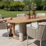 Ein Mann mit Sonnenbrille und Freizeitkleidung sitzt an einem Estate Gartenmöbel-Diningtisch teak natur von 4SO mit sechs beigen Bernini Stühlen auf einer Veranda. Auf dem Tisch in Mangoform stehen zwei Terrakotta-Vasen mit Pflanzen. Grünes Gras und Sonnenlicht füllen den Hintergrund.