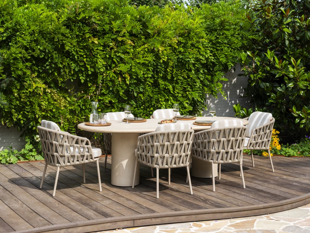 Esstisch im Freien auf einer Holzterrasse mit sechs Flechtstühlen der Serie Beverly von 4SO Dining-Stuhl um einen ovalen Hampton Tisch mit Tellern, Gläsern und einer Wasserkaraffe. Üppiges grünes Laub bildet einen blattreichen Hintergrund, der an einen Garten oder Hinterhof erinnert.