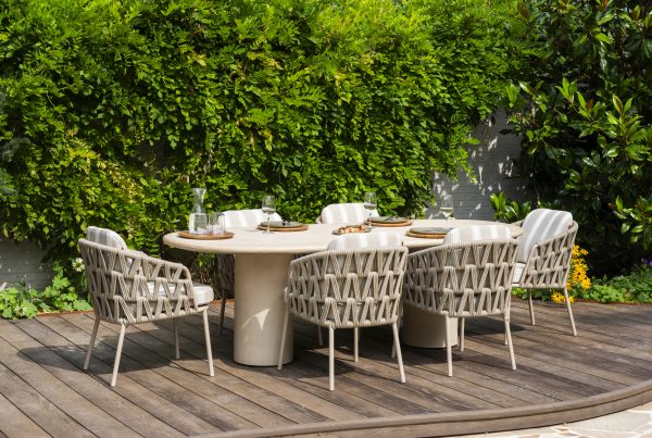 Esstisch im Freien auf einer Holzterrasse mit sechs Flechtstühlen der Serie Beverly von 4SO Dining-Stuhl um einen ovalen Hampton Tisch mit Tellern, Gläsern und einer Wasserkaraffe. Üppiges grünes Laub bildet einen blattreichen Hintergrund, der an einen Garten oder Hinterhof erinnert.