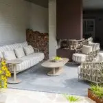 Moderne Außenterrasse mit dem 4SO Beverly Lounge Set mit Boaz Tisch: ein geflochtenes Living-Sofa und zwei Sessel mit gestreiften Kissen. Auf dem Couchtisch aus Stein liegen grüne Äpfel. Brennholz, gelbe Blumen und üppiges Grün vervollständigen den Raum.