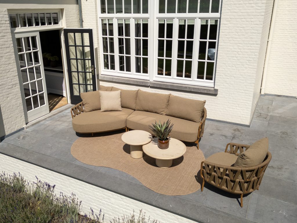 Terrasse im Freien mit einem beigen Sektionssofa, einem passenden Sessel und zwei runden Hampton Couchtischen auf einem hellbraunen Teppich der Serie Biarritz. Der Hamburger Stil der Gartenmöbel wird durch graue Fliesen und ein weißes Backsteinhaus mit großen Fenstern und einer schwarz gerahmten Tür unterstrichen.