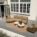 Terrasse im Freien mit einem beigen Sektionssofa, einem passenden Sessel und zwei runden Hampton Couchtischen auf einem hellbraunen Teppich der Serie Biarritz. Der Hamburger Stil der Gartenmöbel wird durch graue Fliesen und ein weißes Backsteinhaus mit großen Fenstern und einer schwarz gerahmten Tür unterstrichen.