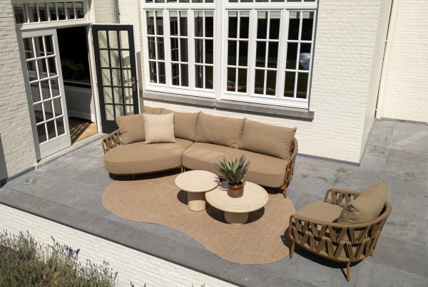 Terrasse im Freien mit einem beigen Sektionssofa, einem passenden Sessel und zwei runden Hampton Couchtischen auf einem hellbraunen Teppich der Serie Biarritz. Der Hamburger Stil der Gartenmöbel wird durch graue Fliesen und ein weißes Backsteinhaus mit großen Fenstern und einer schwarz gerahmten Tür unterstrichen.