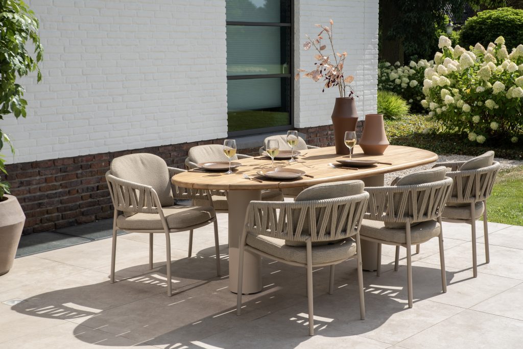 Ein moderner Esstisch im Freien mit sechs gepolsterten Calma Dining-Stühlen von 4SO Gartenmöbeln in Farbe Latte steht auf einer gefliesten Veranda. Der Estate Tisch ist mit Tellern, Weingläsern und braunen Vasen mit Zweigen gedeckt, neben einem weißen Backsteingebäude inmitten von Grün und blühenden Sträuchern.