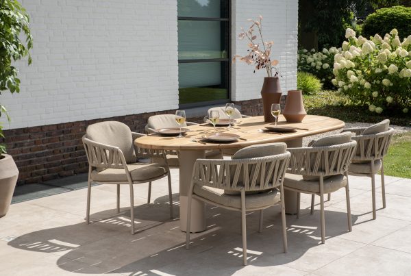 Ein moderner Esstisch im Freien mit sechs gepolsterten Calma Dining-Stühlen von 4SO Gartenmöbeln in Farbe Latte steht auf einer gefliesten Veranda. Der Estate Tisch ist mit Tellern, Weingläsern und braunen Vasen mit Zweigen gedeckt, neben einem weißen Backsteingebäude inmitten von Grün und blühenden Sträuchern.