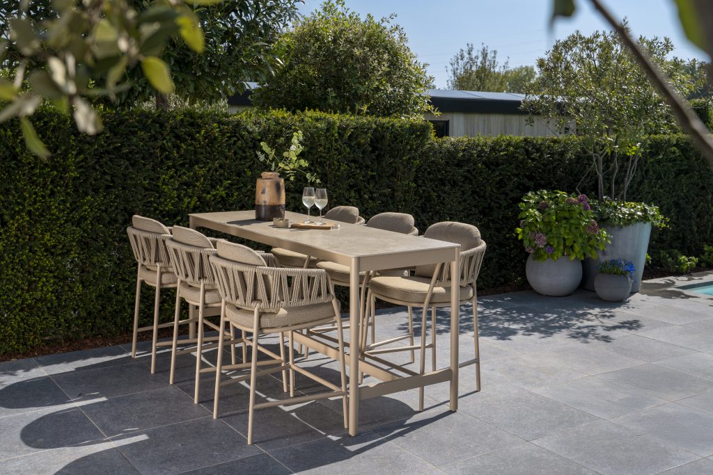 Ein modernes Outdoor-Dining-Set - bestehend aus acht Calma High-Dining-Stühlen und einem passenden Soho High-Diningtisch 220x75x95cm in Keramik Latte von 4SO - steht auf einer Steinterrasse. Üppiges Grün und Topfpflanzen bilden eine einladende Kulisse.