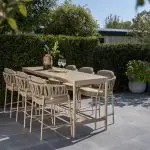 Ein modernes Outdoor-Dining-Set - bestehend aus acht Calma High-Dining-Stühlen und einem passenden Soho High-Diningtisch 220x75x95cm in Keramik Latte von 4SO - steht auf einer Steinterrasse. Üppiges Grün und Topfpflanzen bilden eine einladende Kulisse.