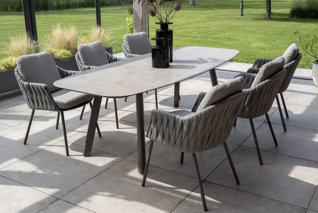 In einem modernen Essbereich im Freien stehen Dining-Stühle aus der Serie Cannes von 4SO in Farbe Terre, die um einen rechteckigen Tisch mit einer steinähnlichen Platte angeordnet sind. Zwei hohe schwarze Vasen vervollständigen den Look auf einer gefliesten Terrasse mit Blick auf einen grünen Rasen und Garten.