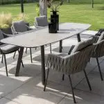 In einem modernen Essbereich im Freien stehen Dining-Stühle aus der Serie Cannes von 4SO in Farbe Terre, die um einen rechteckigen Tisch mit einer steinähnlichen Platte angeordnet sind. Zwei hohe schwarze Vasen vervollständigen den Look auf einer gefliesten Terrasse mit Blick auf einen grünen Rasen und Garten.