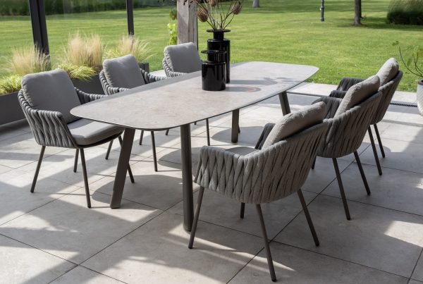 In einem modernen Essbereich im Freien stehen Dining-Stühle aus der Serie Cannes von 4SO in Farbe Terre, die um einen rechteckigen Tisch mit einer steinähnlichen Platte angeordnet sind. Zwei hohe schwarze Vasen vervollständigen den Look auf einer gefliesten Terrasse mit Blick auf einen grünen Rasen und Garten.