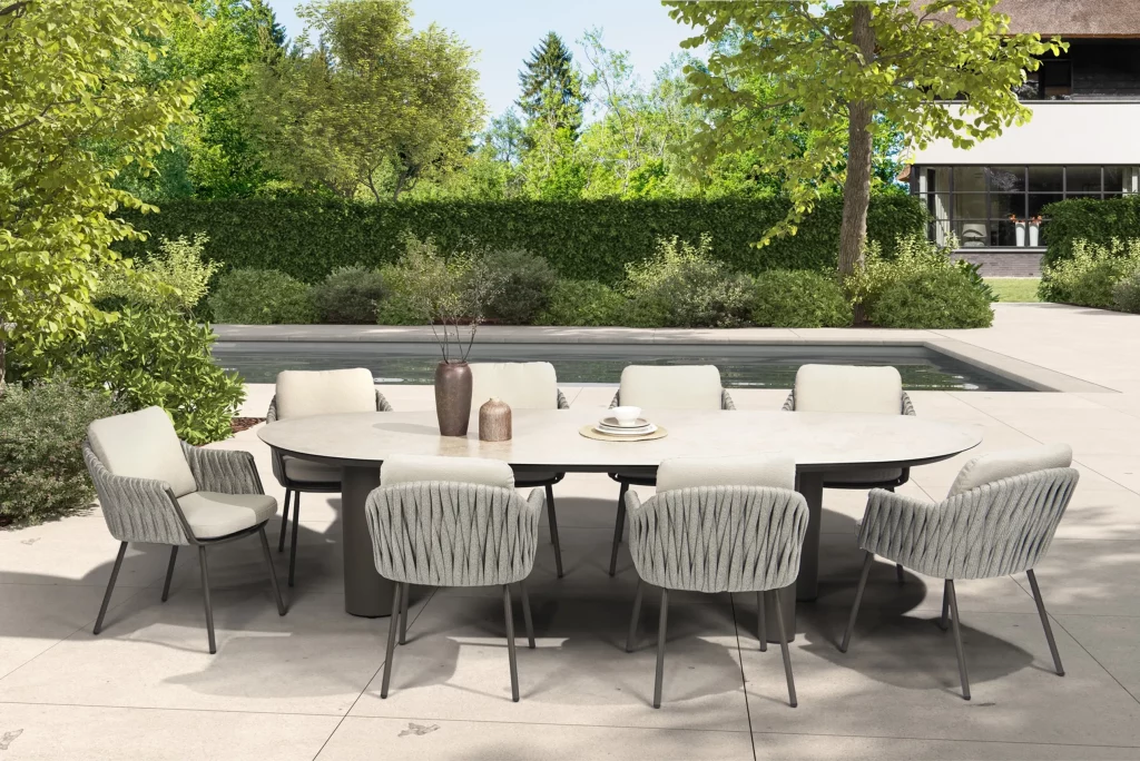 Outdoor-Essgarnitur auf einer Terrasse mit acht gepolsterten Stühlen um einen Denia Diningtisch Oval 290x110cm mit Keramikdruck Latte. Der Tisch ist mit Vasen und Tellern dekoriert, mit Blick auf einen Pool, Gartenmöbel, grüne Sträucher und ein modernes Haus unter einem hellen Himmel.