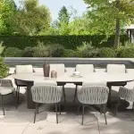 Outdoor-Essgarnitur auf einer Terrasse mit acht gepolsterten Stühlen um einen Denia Diningtisch Oval 290x110cm mit Keramikdruck Latte. Der Tisch ist mit Vasen und Tellern dekoriert, mit Blick auf einen Pool, Gartenmöbel, grüne Sträucher und ein modernes Haus unter einem hellen Himmel.