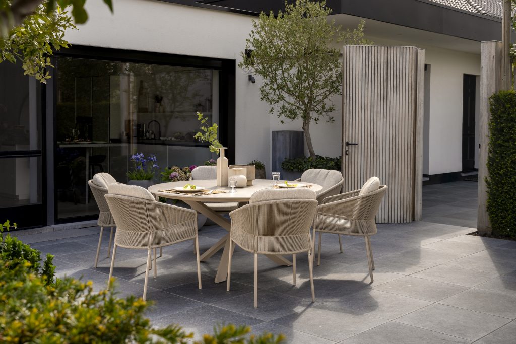 Eine moderne Außenterrasse mit Gartenmöbeln der Serie Florence von 4SO in Farbe Latte, mit einem runden Prado Holztisch für sechs Personen, gepolsterten Stühlen, grauem Fliesenboden, Topfpflanzen und einem weißen Gebäude mit großen Fenstern und einer gerippten Holztür.