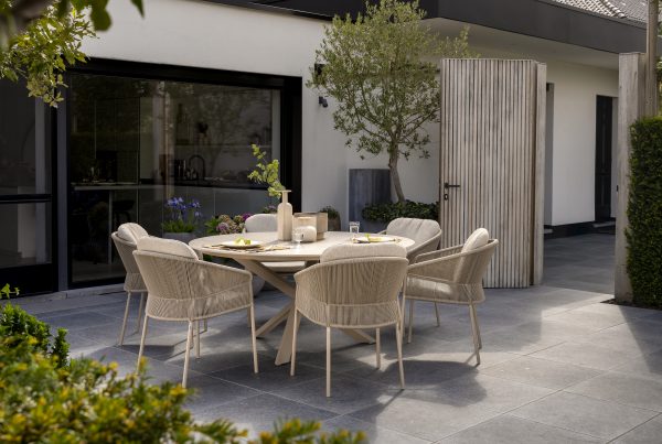 Eine moderne Außenterrasse mit Gartenmöbeln der Serie Florence von 4SO in Farbe Latte, mit einem runden Prado Holztisch für sechs Personen, gepolsterten Stühlen, grauem Fliesenboden, Topfpflanzen und einem weißen Gebäude mit großen Fenstern und einer gerippten Holztür.
