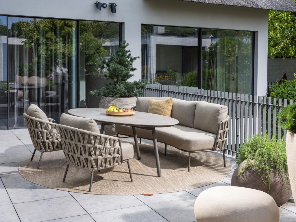 Moderne Außenterrasse mit beigefarbenen gepolsterten Dining-Stühlen und Sofa aus der Serie Mallorca von 4SO, drei passenden geflochtenen Sesseln und einem ovalen Salute Tisch mit einer Obstschale. Die Sitzgruppe steht auf einem runden Teppich inmitten von Topfpflanzen und großen Glasfenstern.