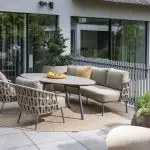 Moderne Außenterrasse mit beigefarbenen gepolsterten Dining-Stühlen und Sofa aus der Serie Mallorca von 4SO, drei passenden geflochtenen Sesseln und einem ovalen Salute Tisch mit einer Obstschale. Die Sitzgruppe steht auf einem runden Teppich inmitten von Topfpflanzen und großen Glasfenstern.