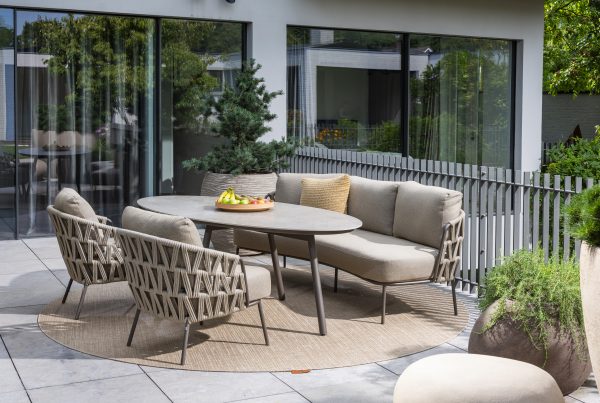 Moderne Außenterrasse mit beigefarbenen gepolsterten Dining-Stühlen und Sofa aus der Serie Mallorca von 4SO, drei passenden geflochtenen Sesseln und einem ovalen Salute Tisch mit einer Obstschale. Die Sitzgruppe steht auf einem runden Teppich inmitten von Topfpflanzen und großen Glasfenstern.