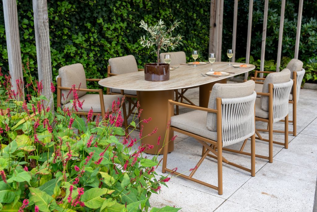 Ein Outdoor-Dining-Set aus der Serie Marina von 4SO mit sechs gepolsterten Dining-Stühlen und einem ovalen Tisch steht auf einer Steinterrasse. Auf dem Colorado Tisch stehen eine Topfpflanze, drei Gläser mit bernsteinfarbenem Wein und Teller mit Speisen inmitten von üppigem Grün und roten Blumen.
