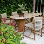 Ein Outdoor-Dining-Set aus der Serie Marina von 4SO mit sechs gepolsterten Dining-Stühlen und einem ovalen Tisch steht auf einer Steinterrasse. Auf dem Colorado Tisch stehen eine Topfpflanze, drei Gläser mit bernsteinfarbenem Wein und Teller mit Speisen inmitten von üppigem Grün und roten Blumen.