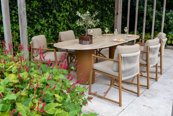 Ein Outdoor-Dining-Set aus der Serie Marina von 4SO mit sechs gepolsterten Dining-Stühlen und einem ovalen Tisch steht auf einer Steinterrasse. Auf dem Colorado Tisch stehen eine Topfpflanze, drei Gläser mit bernsteinfarbenem Wein und Teller mit Speisen inmitten von üppigem Grün und roten Blumen.